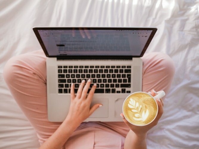 A lady in her pajamas working from home. Typing on her laptop and holding a mug of coffee