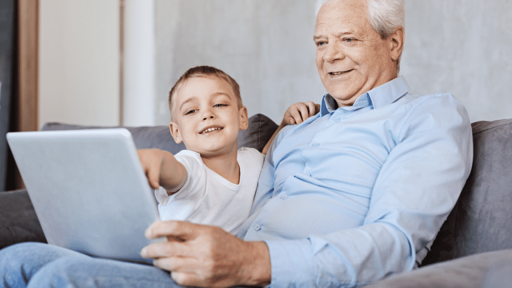 A grandfather and his grandson sitting on the sofa and grandson pointing at the laptop screen
